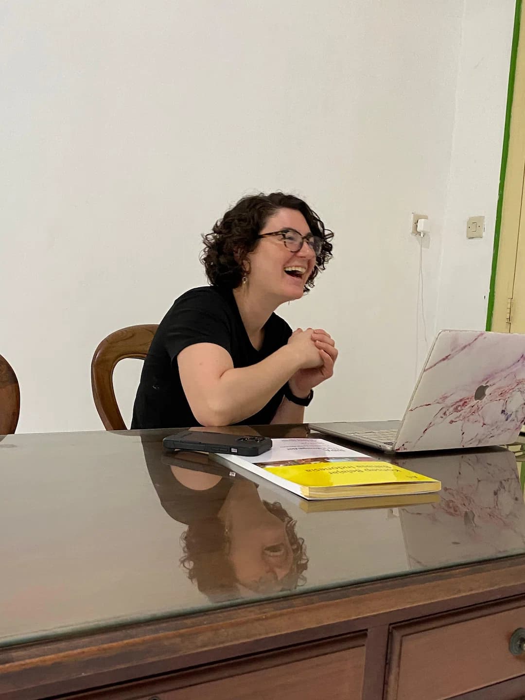 A Puri Bahasa student laughing while studying at a desk with a laptop and textbook